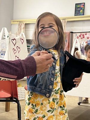 A student smiles behind a magnifying glass, enlarging her smile